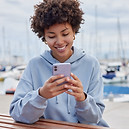 young-cheerful-curly-woman-uses-mobile-phone-sits-pier-near-sea-reads-pleasant-text-messag
