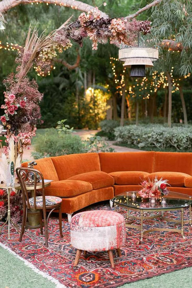 Cozy outdoor lounge with orange sofa, patterned rug, floral decor, and string lights. A glass table holds a flower arrangement.