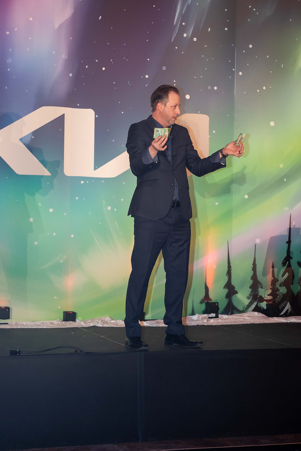 A person in a suit holds money on stage against a colorful backdrop. The setting features Aurora Borealis-like patterns and a large "KIA" logo.