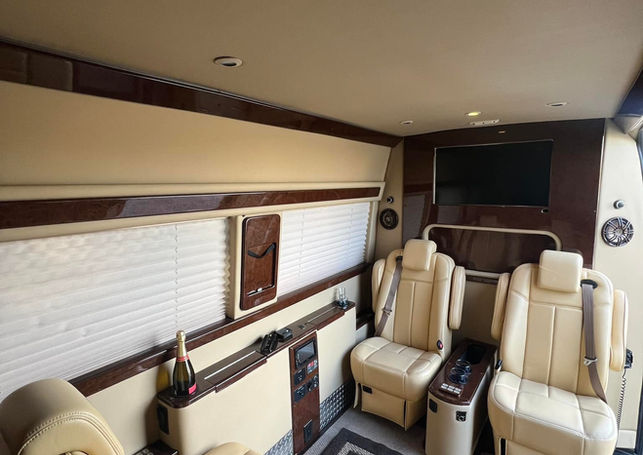 A luxury van interior with cream leather seating, a mounted TV, and wood trim.