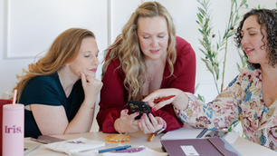 Three women collaborate at a table, focused on a smartphone. One points at the screen. Background shows plants; mood is engaged.