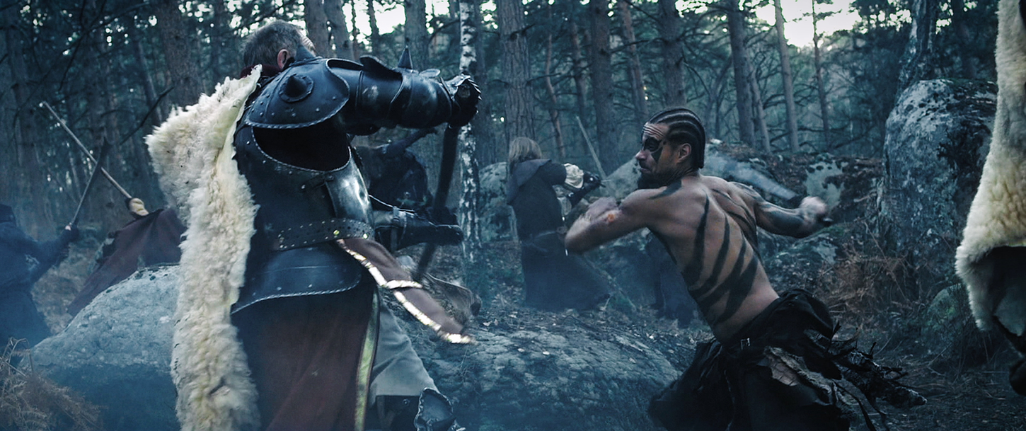 Dans une sombre forêt, un homme âgé en armure se bat contre un homme tatoué et mutilé.