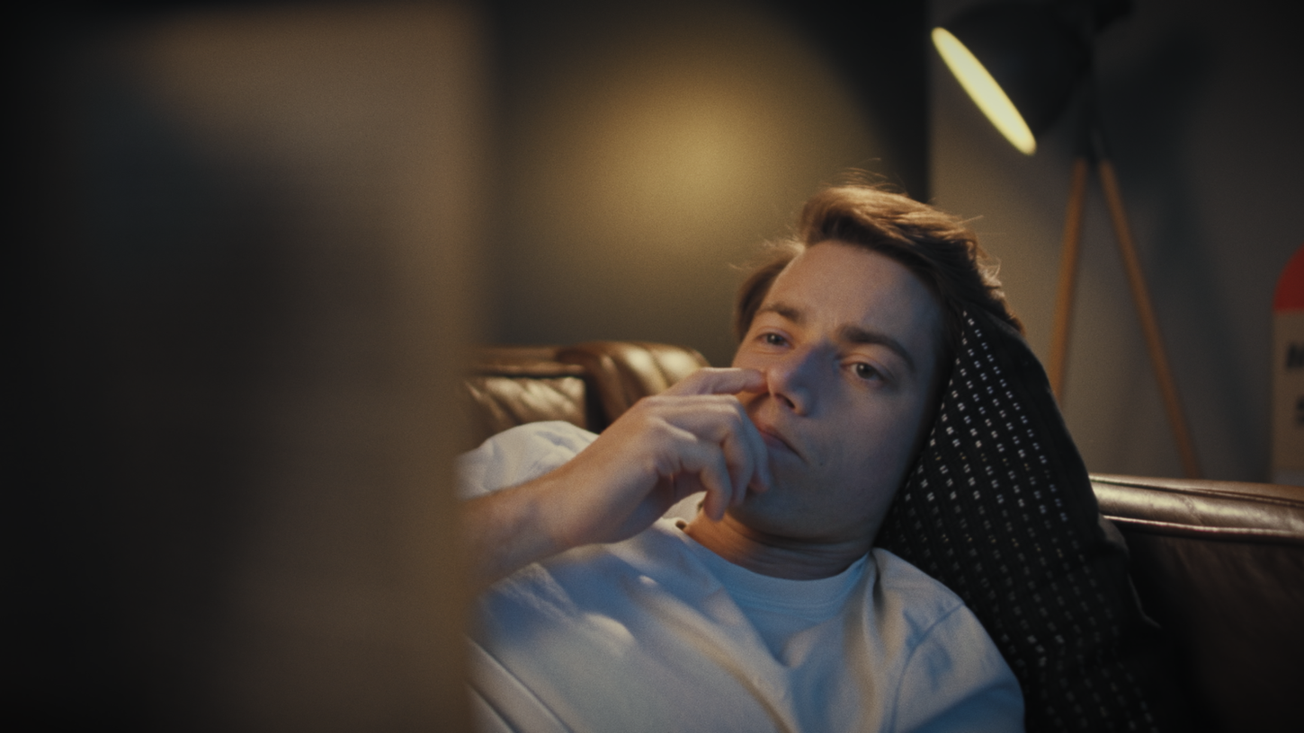 Un jeune homme met son doigt dans son nez en lisant un livre, allongé sur un canapé.