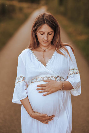 Femme enceinte en robe blanche pose de face et les yeux tournées vers son enfant à naître sur une route de campagne à Villars les Dombes un soir au soleil couchant