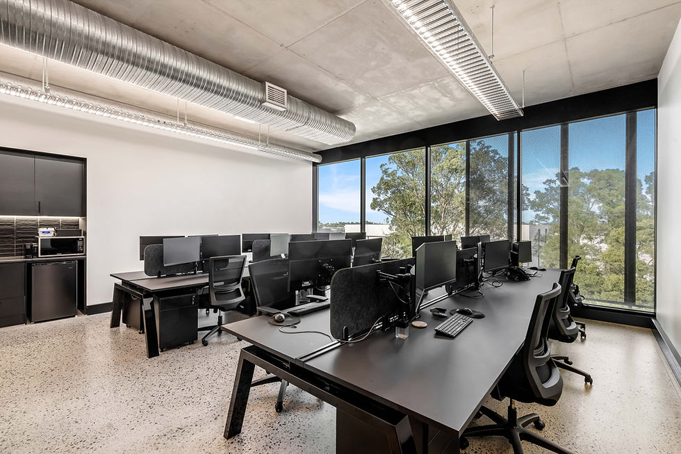 Modern office with black desks and computers, empty chairs. Large windows reveal trees outside. Bright, industrial feel with concrete ceiling.