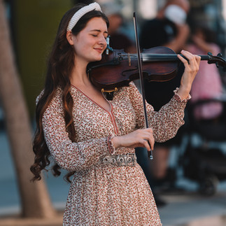Avelina Kushnir playing violin on the street in Santa Monica, eyes closed, smiling in a floral dress