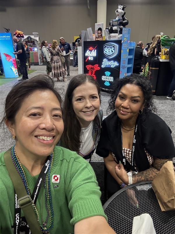 Three women smiling at a convention, sitting by a table. Colorful signs in the background. Lanyards and badge details visible.