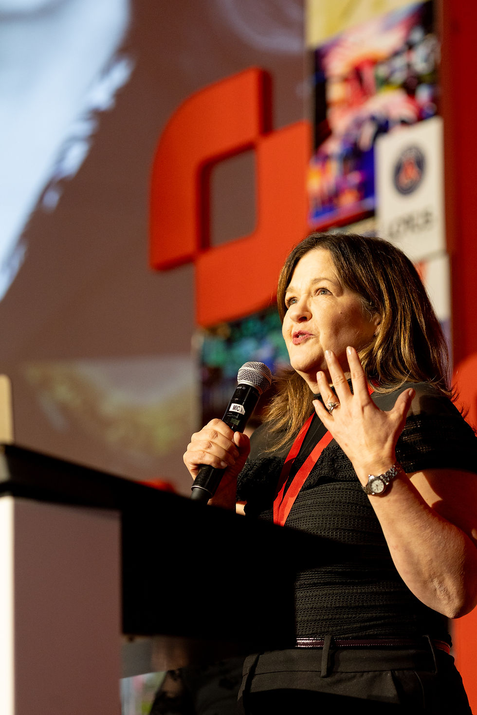 Helen Herrick speaking into a microphone onstage, gesturing beside a red logo backdrop at a conference, with a focused expression.