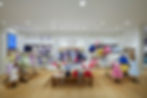 Janie and Jack clothing store with vibrant dresses on racks and folded clothes on a table. Brightly lit, with framed images on the back wall.