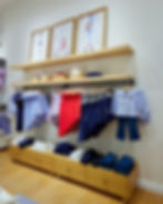 Janie and Jack clothing store display with colorful dresses and shirts on hangers. Wooden shelves have framed photos above folded clothes.