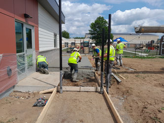 Five construction workers pouring new concrete pathway beside orange building.