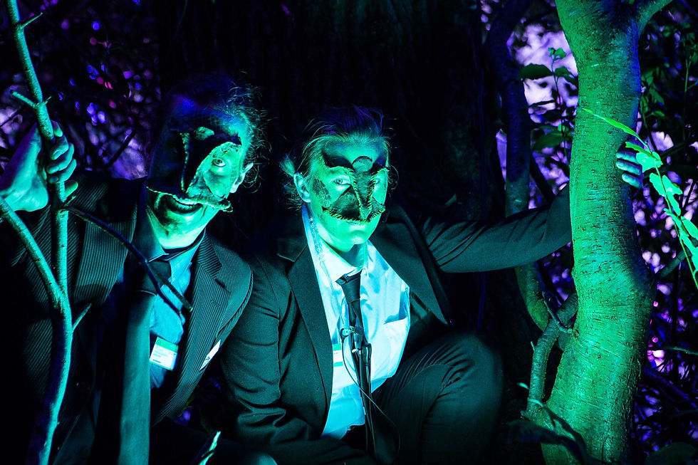 2 actors with Lizard face prosthetics, green face paint and suits in a woodland at night