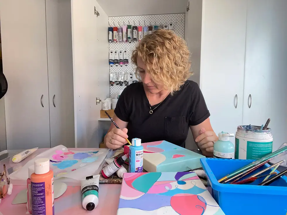 Sandra Vincent paints at a table in a studio, surrounded by art supplies. Bright abstract artwork is visible. Calm and focused mood.
