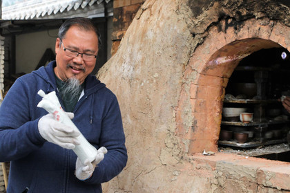 Cheah Yeow Seng holding his small ceramic lady sculptor artwork in front of a pottery wood kiln in winter wear.