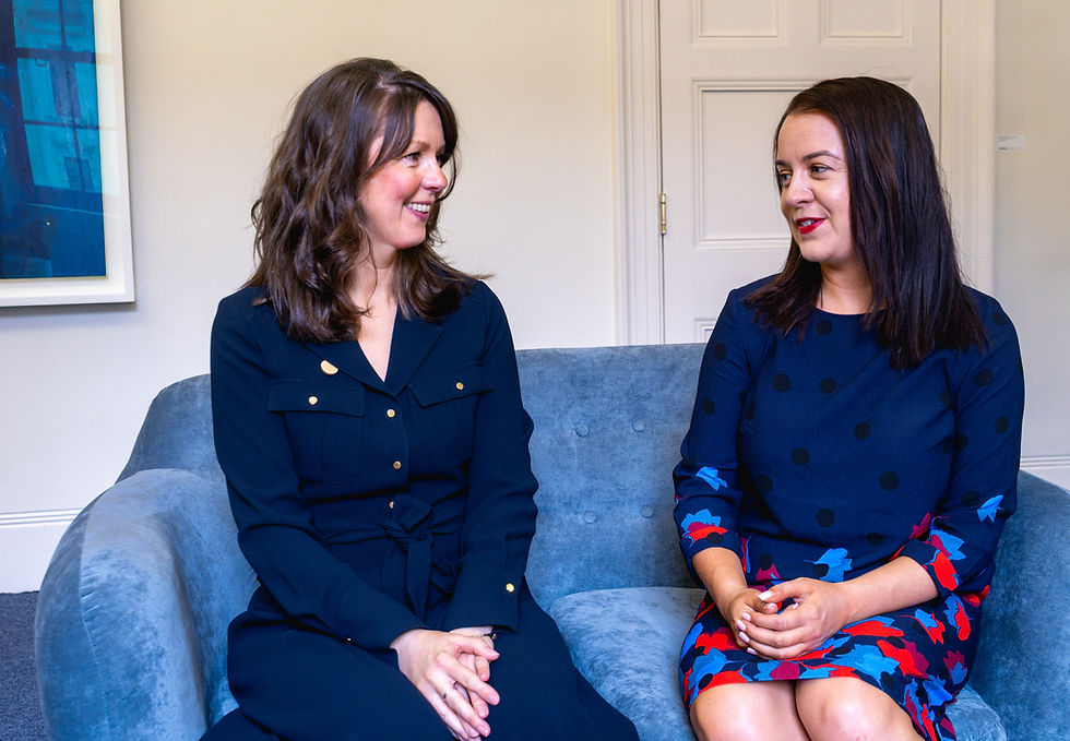 Amy and Stephanie sit smiling at one another on a blue sofa. Amy is wearing a blue dress with brass buttons, while Stephanie wears a blue polka dot dress and a blue and red skirt.