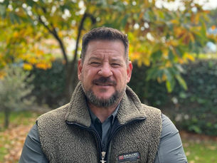 A man stands outdoors in an autumnal setting, wearing a cozy fleece vest with a hint of a smile. The background is filled with vibrant fall foliage, adding warmth and color to the scene.