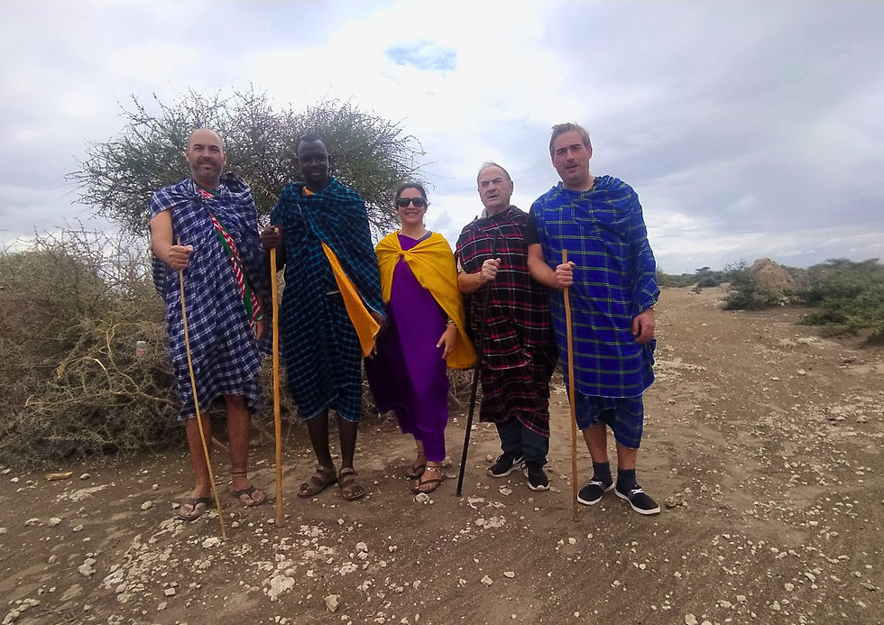 Cinco personas con túnicas maasai coloridas y bastones posan sonrientes en un paisaje árido con matorrales. El cielo está parcialmente nublado.
