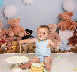Photo of 1 year old boy in blue romper smiling and holding spoon after knocking his cake off the stand in 1st Birthday Cake Smash Photoshoot East Yorkshire, Hull and North Lincolnshire.