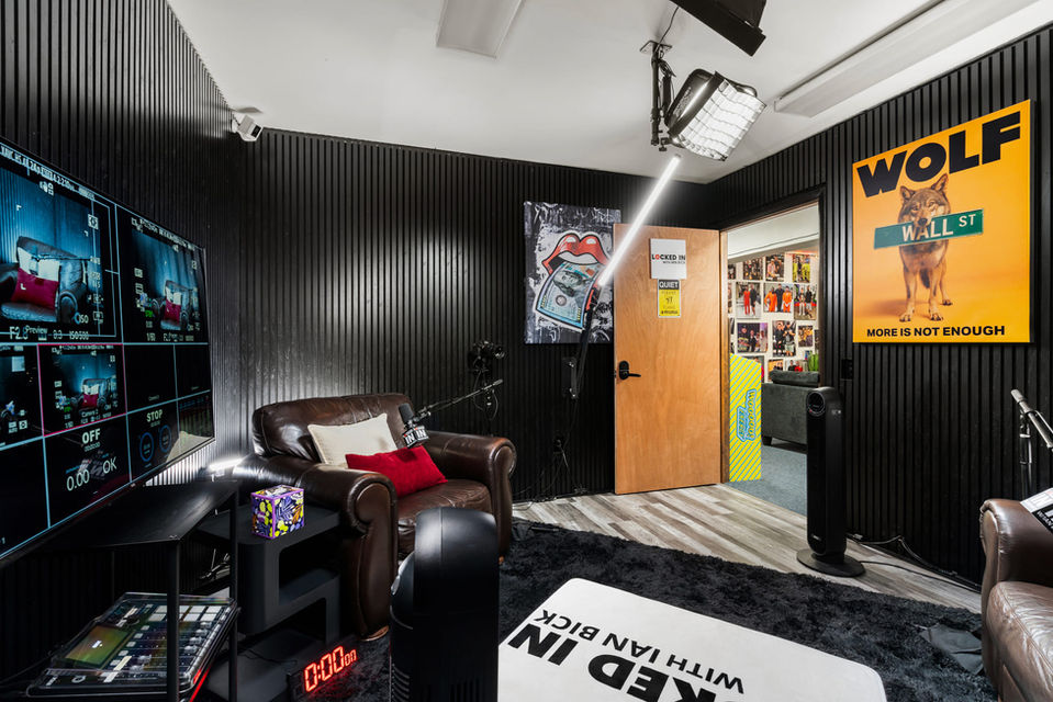 Side angle of the Locked In podcast studio showing camera rigs, LED lights, control monitors, leather chairs, and black acoustic wall panels.