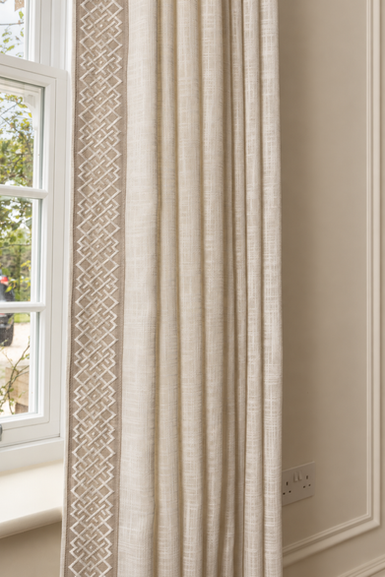 Close-up of cream textured curtains with an embroidered geometric border beside a sash window.