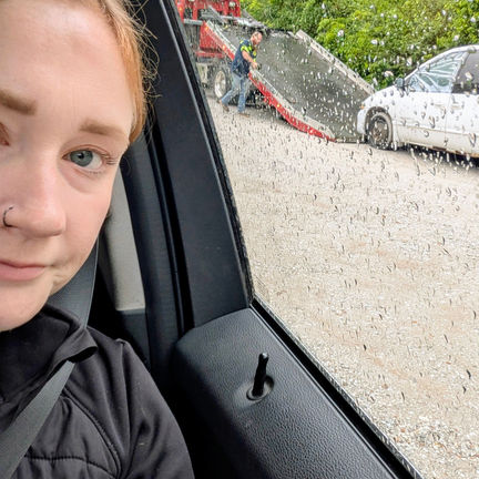 A tired woman taking a selfie as a vehicle is being towed in the background