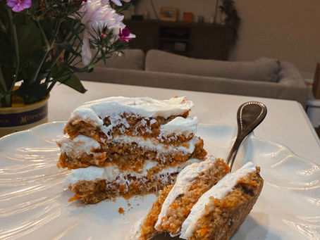 Carrot cake with white frosting on a plate with a fork, set on a table. Colorful flowers in the background, giving a cozy vibe.