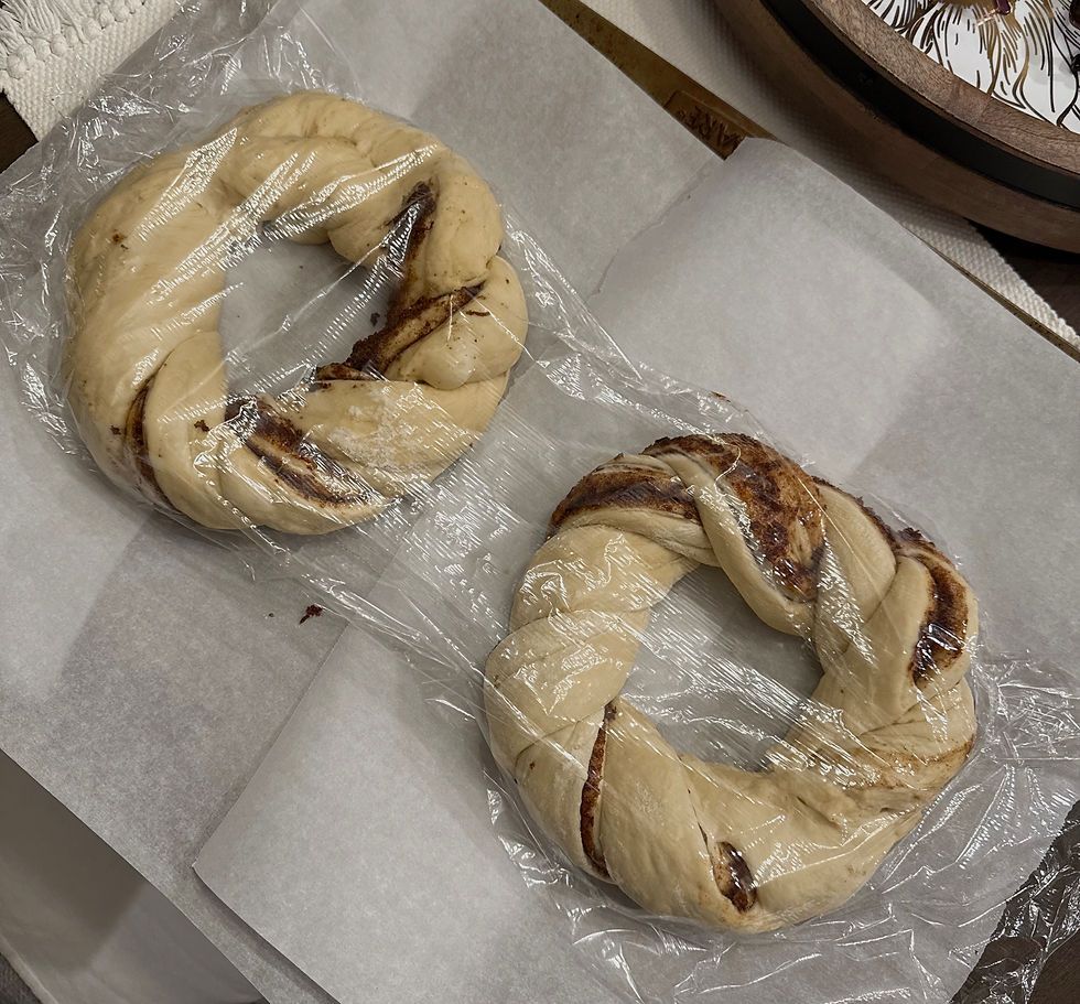 Two uncooked braided dough wreaths with cinnamon swirls, wrapped in plastic on parchment paper, on a kitchen counter.
