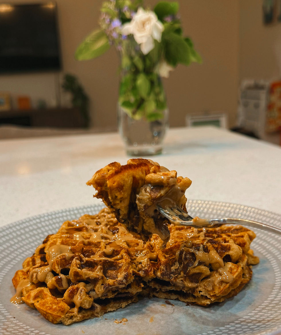Sweet potato sourdough discard waffles on a plate topped with syrup, with a fork lifting a bite. A vase of flowers in the blurred background on a kitchen table.