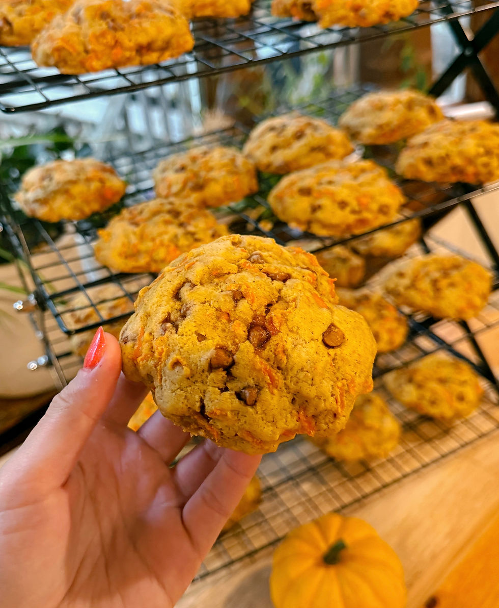 Hand holding a carrot cake cookie with visible cinnamon chips, in front of a cooling rack of similar cookies. A small pumpkin decorates the table.