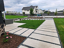 A stunning step-stone walkway leading to a pool surrounded by young trees, and green grass separated by hardscape walks and patios.