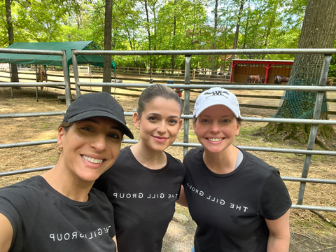 Maral, Gina, and Alison from The Gill Group smiling outdoors in matching black "The Gill Group" T-shirts during KW Red Day, a Keller Williams community service event, with a sunny wooded backdrop and metal fence.