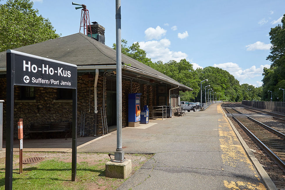 Ho-Ho-Kus train station sign and tracks under a clear sky. Stone building nearby, surrounded by greenery.