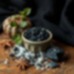 Ceramic bowl of black salt on wooden surface with star anise, scattered salt, black stones, and a small pumpkin, against a dark cloth backdrop.