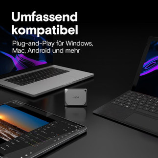 Eine graue portable Crucial SSD zentral positioniert zwischen einem geöffneten Laptop, einem Tablet mit Bildbearbeitungsprogramm und einem weiteren Laptop oder Tablet mit Tastatur. Der Hintergrund ist dunkel mit lila und blauen Akzenten. Sichtbarer Text 'Umfassend kompatibel' und 'Plug-and-Play für Windows, Mac, Android und mehr'.