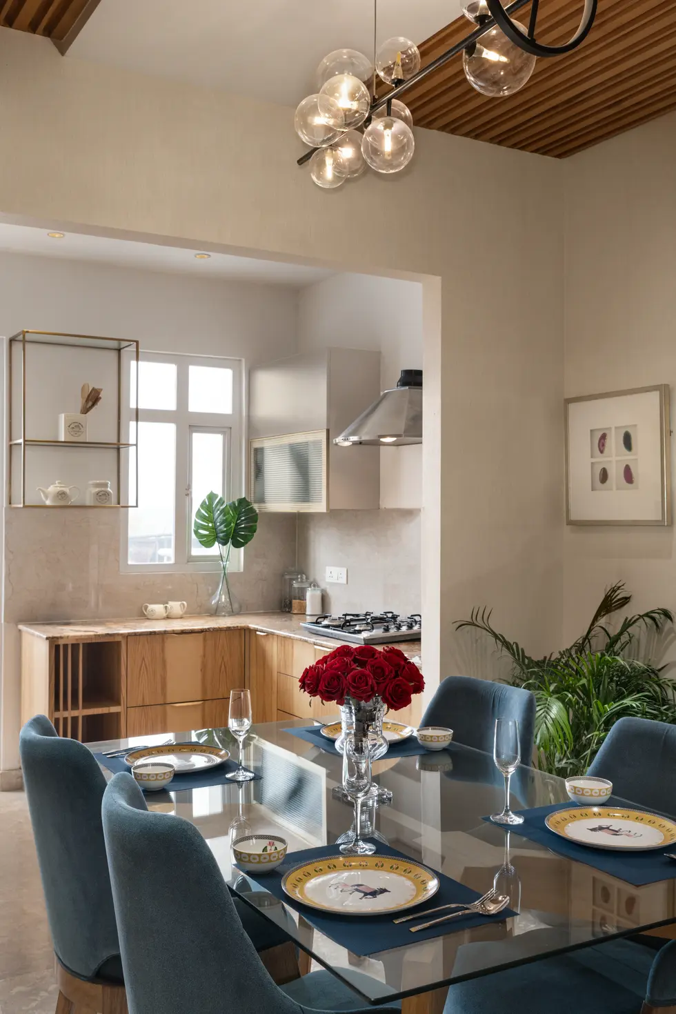 Modern dining area with blue upholstered chairs and glass table, connected to a semi-open modular kitchen with light wood cab