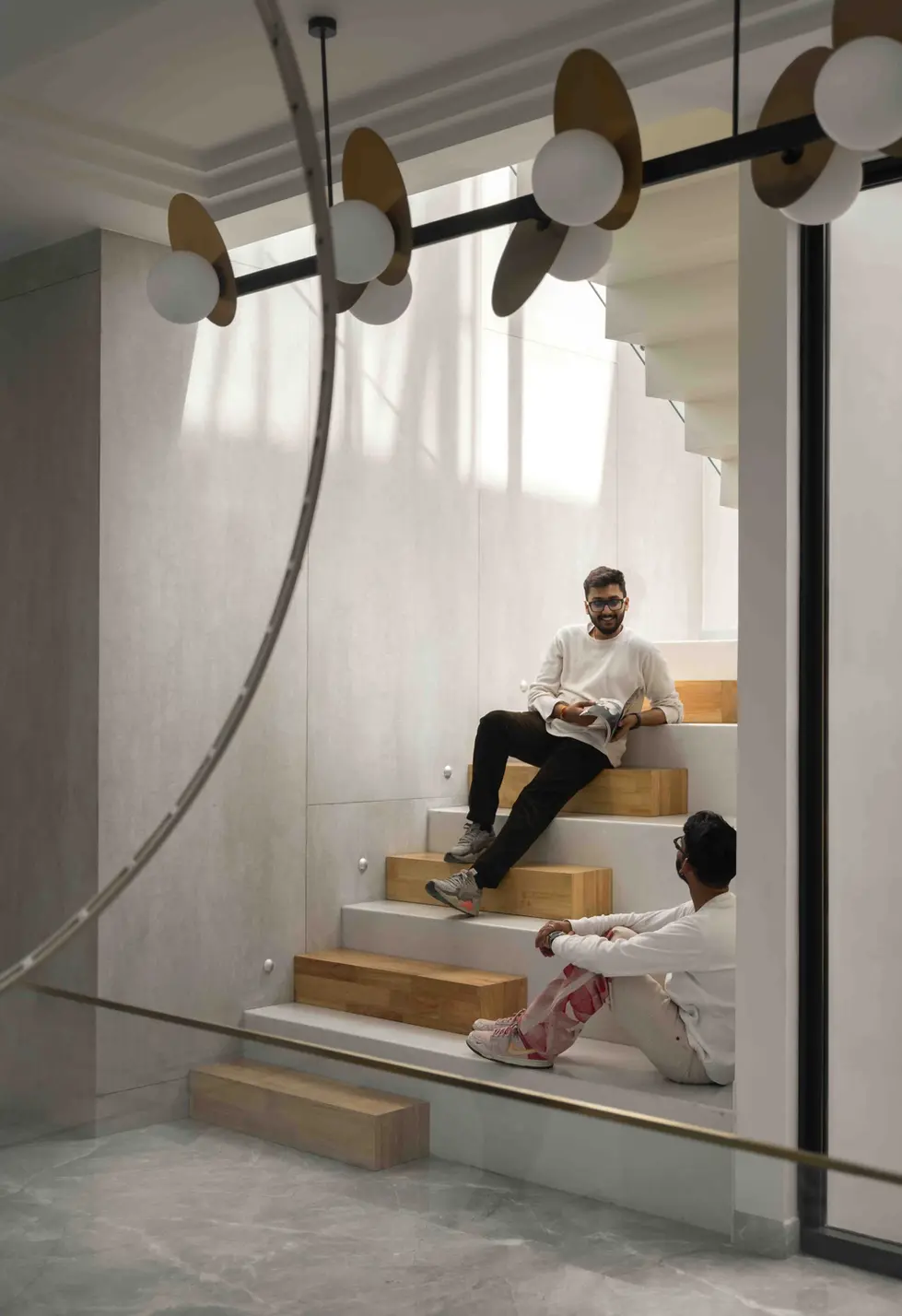 Double-height atrium staircase illuminated by a skylight, featuring white marble steps, glass railings, and large sculptural bronze ring chandeliers suspended from the ceiling.