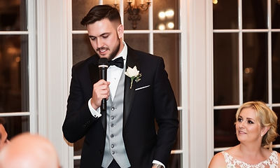 a groomsman is speaking on the microphone at a wedding dinner.