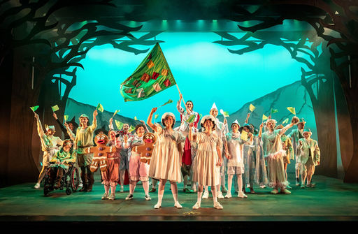 On a fairytale-inspired stage, a vibrant ensemble of fairytale creatures stands united, with two performers in matching white outfits and eggshell-like hats taking center stage while another stands tall in the center background holding a large, colorful patchwork flag. The surrounding cast members wave small green flags, framed by dark, silhouetted forest trees and a brilliant, glowing turquoise backdrop that amplifies the scene's triumphant and whimsical atmosphere.