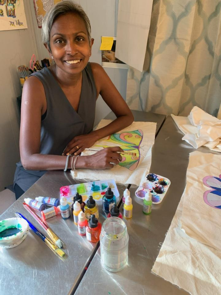 Rachel looks at the camera, smiling. She is sitting at a table with brushes, paints, and an in-progress painting of a butterfly