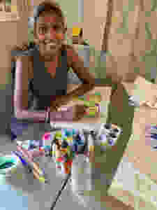 Rachel looks at the camera, smiling. She is sitting at a table with brushes, paints, and an in-progress painting of a butterfly