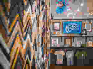 A wall of framing material samples on the left; in the distance are multiple shelves with examples of artwork, and tshirts for sale