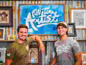 Nathan and Micah smile for the camera in front of a sign that says "Full Time Artist".