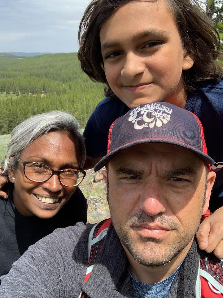 Rachel smiles at the camera, and her son Prakash rests his head on top of Ryan's head. Ryan is squinting as he takes the selfie because the sun is in his eyes. In the background there is a forest of evergreen trees in the distance.