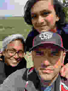 Rachel smiles at the camera, and her son Prakash rests his head on top of Ryan's head. Ryan is squinting as he takes the selfie because the sun is in his eyes. In the background there is a forest of evergreen trees in the distance.