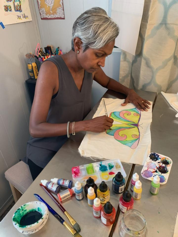 Rachel sits at her art table with brushes, inks, paint, and a palette. She is painting a butterfly on a piece of cotton fabric.