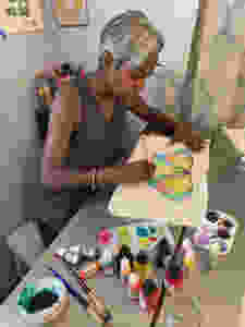 Rachel sits at her art table with brushes, inks, paint, and a palette. She is painting a butterfly on a piece of cotton fabric.