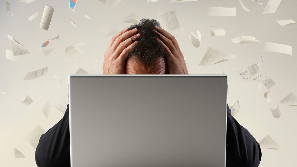 Um empresário ou dona de loja, sentado na frente do computador, olhando para o celular com cara de frustração.