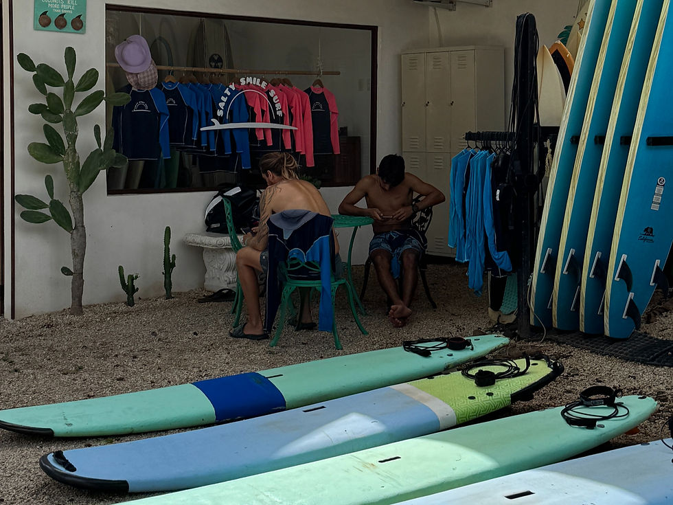 The outdoor area of Salty Smile Surf School in Tamarindo, featuring surfboards laid out on the ground, a rack of standing boards on the right, a shop window displaying colorful rash guards, and two students sitting under a shaded patio surrounded by surf gear and tropical cacti