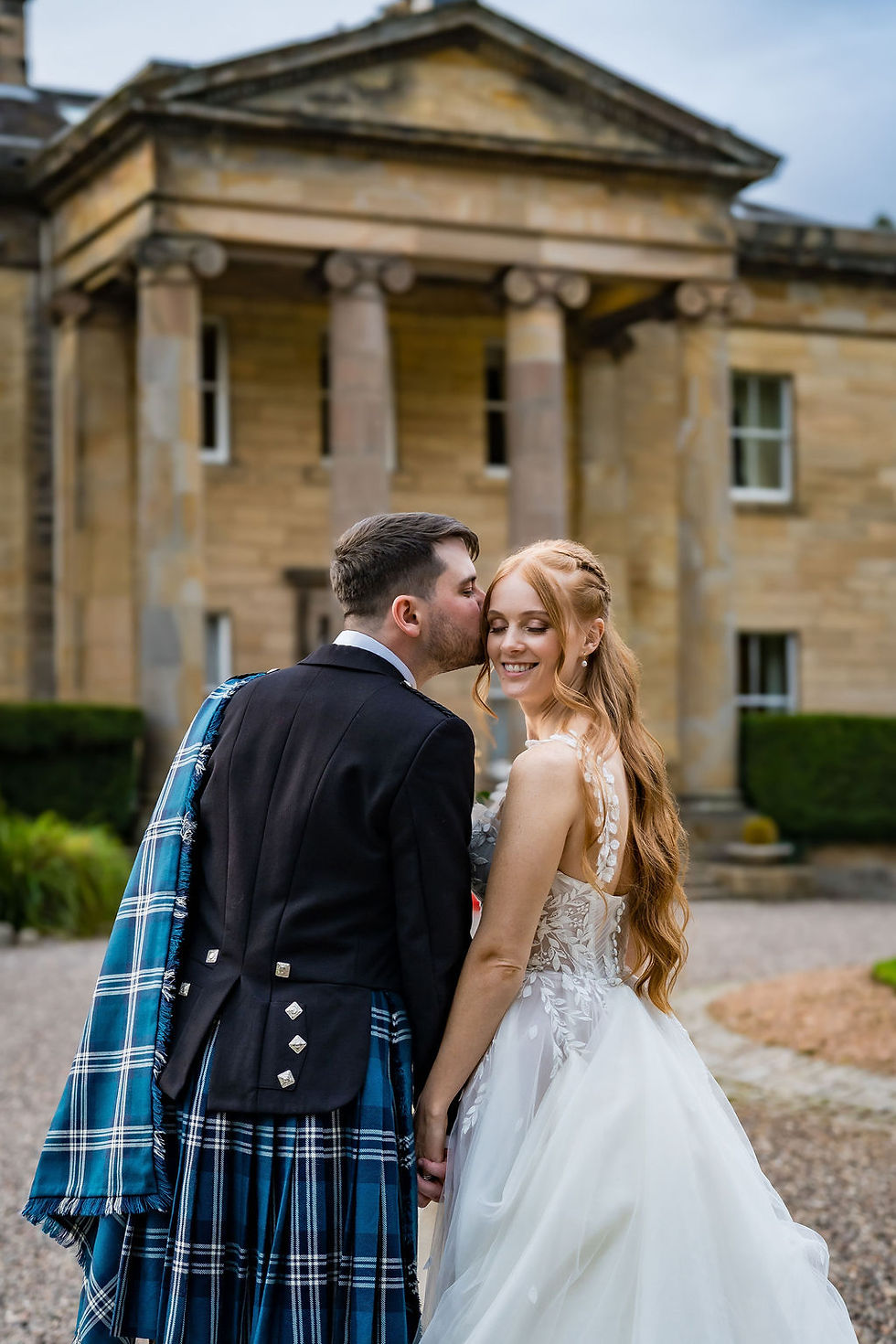 Groom gently kissing his bride on the temple in front of Balbirnie House in Fife, a romantic and natural wedding moment captured by Scottish wedding photographer ZibiZ Photo.