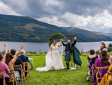 Outdoor wedding ceremony in Scotland where the couple has just been announced as husband and wife, cheering with raised hands as guests celebrate around them, captured in a candid documentary style by a Scottish wedding photographer.